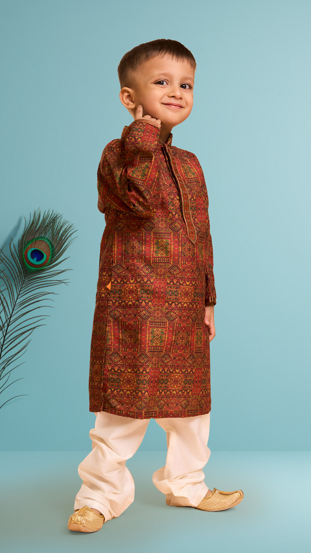 Young boy dressed in a red printed kurta pajama with golden mojaris, posing confidently with a peacock feather in the background for Janmashtami celebration.